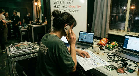 A manager speaks into a phone behind a concert stage, holding a crumpled setlist amidst glowing green stage lights.