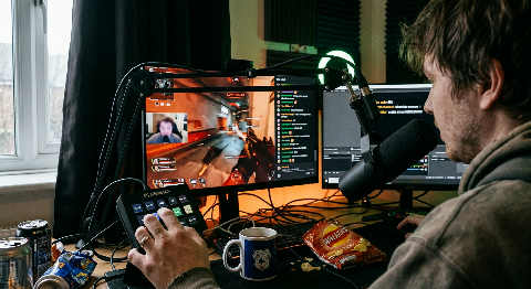 A content creator focuses on three monitors in a dark room, illuminated by the colorful glow of a gaming PC and mic.