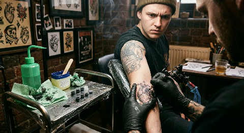 Gloved hands focus intently while guiding a tattoo machine over an arm, ink staining the skin under bright studio lights.