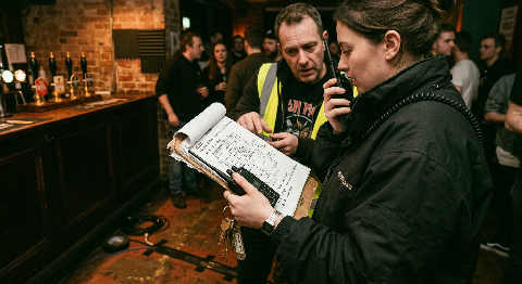 A person in a dark green vest checks a clipboard under moody theater lights while staff blur in the busy background.