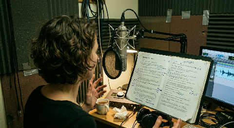 A person speaks into a professional microphone with a pop filter in a dimly lit, soundproofed recording booth.